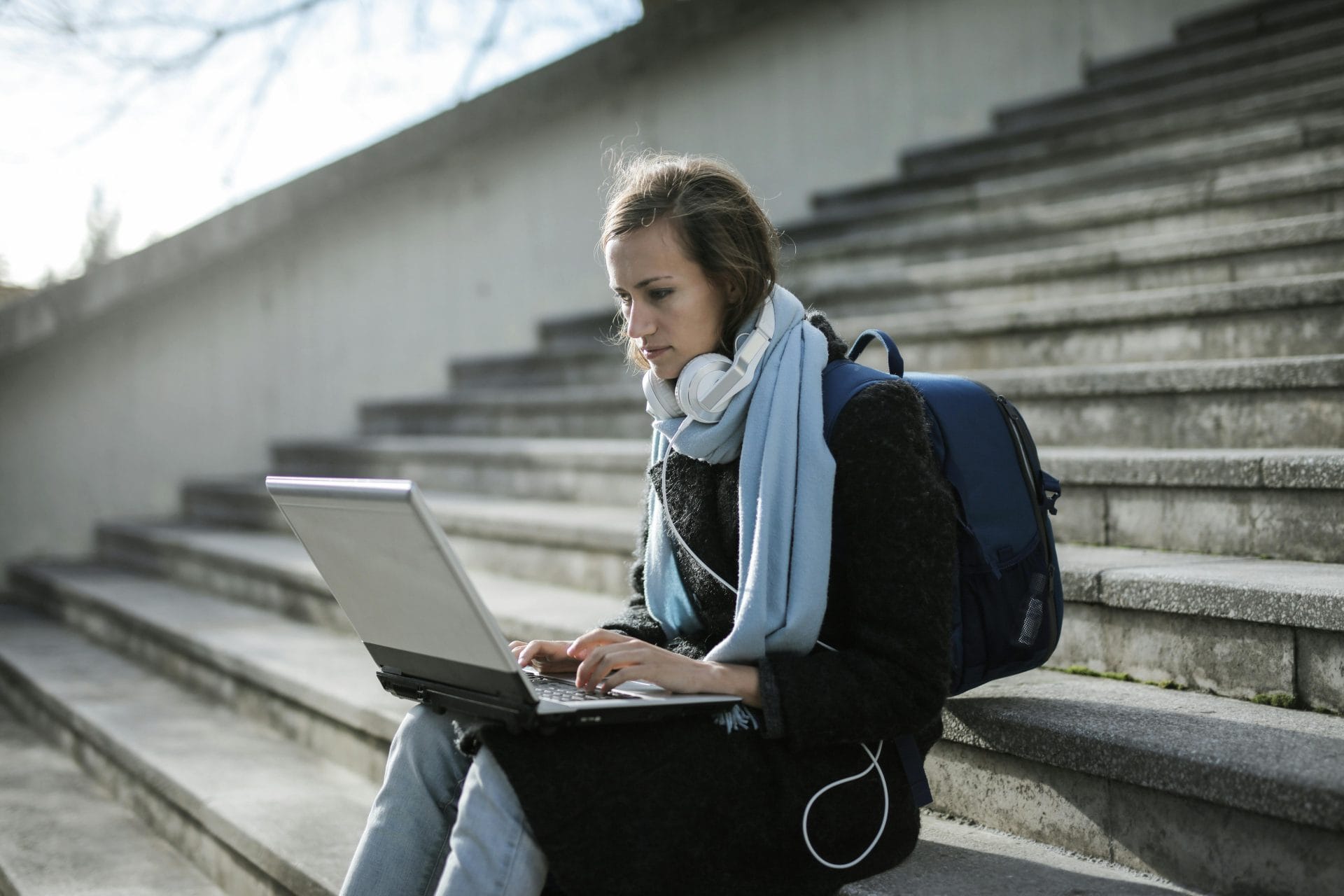 Woman using laptop on outdoor stairs, studying online with headphones and backpack.